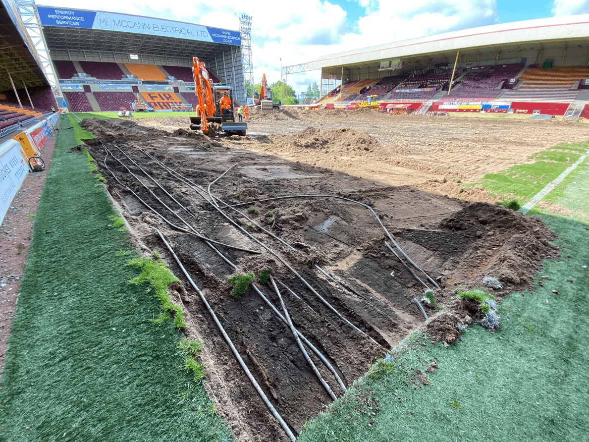Exposed pipework and excavated ground along the edge of the stadium pitch